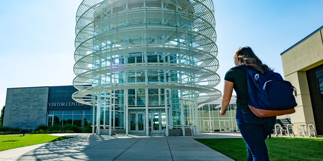 A student wearing a backpack walks toward the entrance of the Visitor Center and University Bookstore.