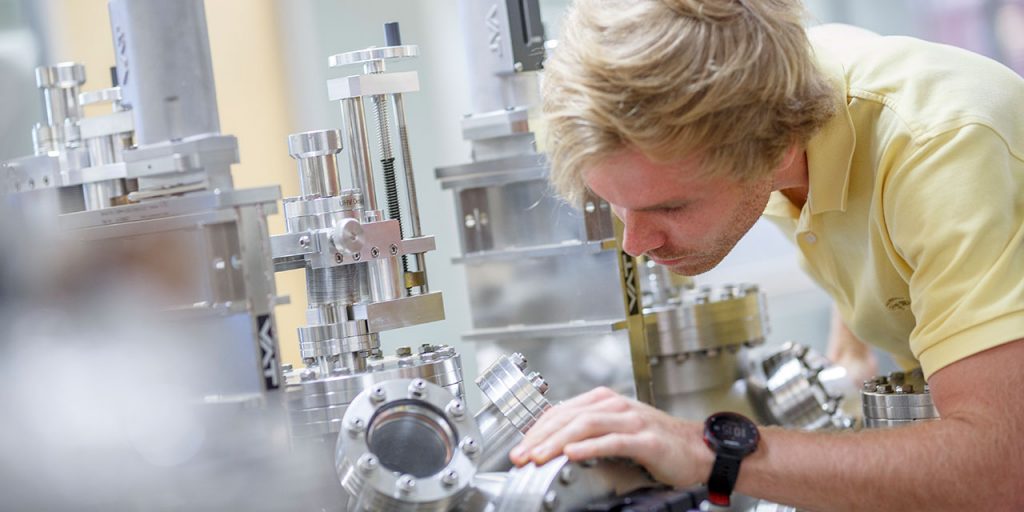 An engineering student in a lab peers into a small window in a large metal device. 
