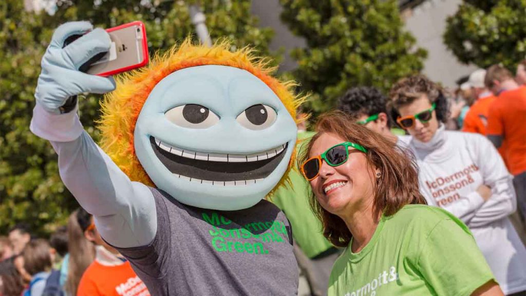 A staff member in orange and green sunglasses poses for a selfie with UTD mascot Temoc.