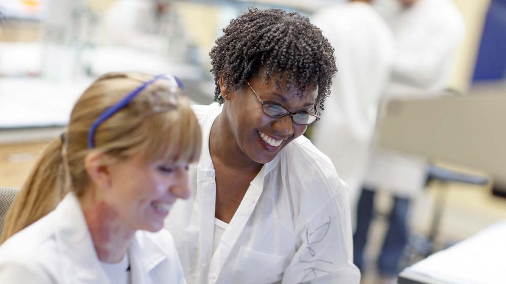Two faculty members in lab coats, with other researchers out of focus in background. 