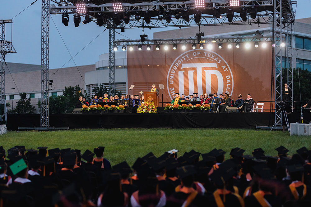 A crowd of graduates listen to the commencement speaker standing on a stage.